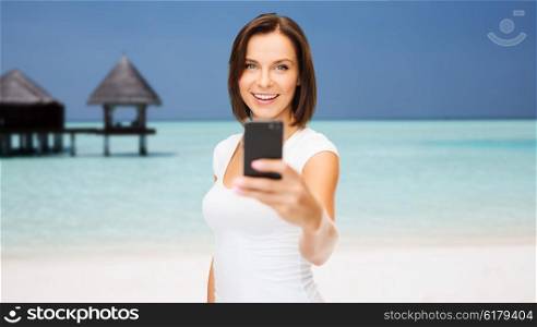 travel, tourism, summer vacation, technology and people - happy young woman taking selfie picture by smartphone on maldives beach with bungalow background