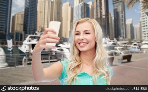 travel, tourism, technology and people concept - happy smiling young woman or teenage girl taking selfie with smartphone over boats in harbor in dubai city background