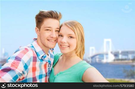 travel, tourism, technology, people and love concept - smiling couple with smartphone or camera taking selfie over rainbow bridge in tokyo and river background