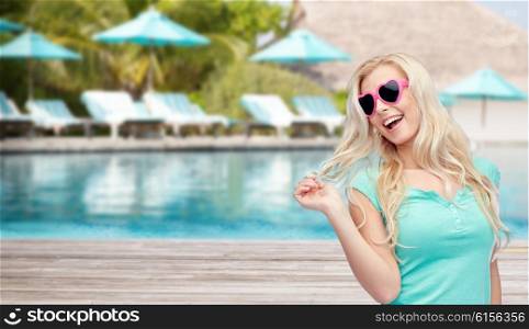 travel, tourism, vacation and people concept - smiling young woman or teenage girl in sunglasses holding her strand of hair over swimming pool on beach with sunbeds and parasol background