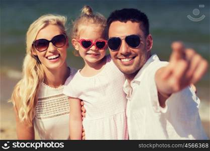 travel, vacation, adoption and people concept - happy family with little girl in sunglasses pointing finger on summer beach