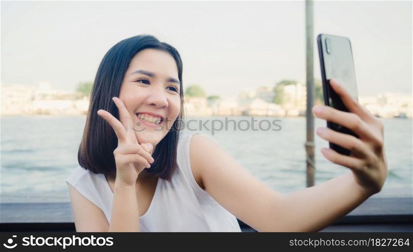 Traveler Asian blogger woman clicking selfie using mobile at cafe, beautiful female using smart phone make vlog and live in social media while enjoying the journey holiday travel near river in city.