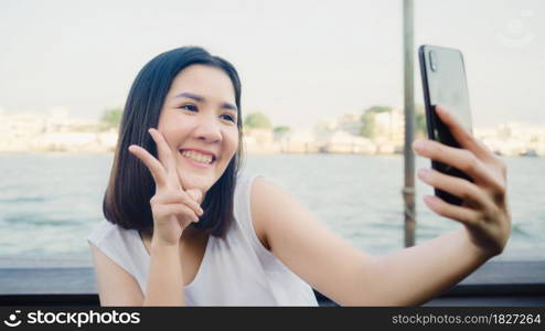 Traveler Asian blogger woman clicking selfie using mobile at cafe, beautiful female using smart phone make vlog and live in social media while enjoying the journey holiday travel near river in city.