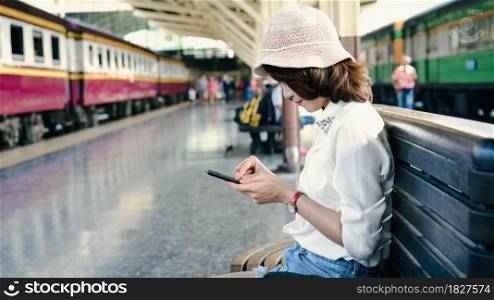 Traveler backpacker Asian woman travel in Bangkok, Thailand. Happy young female sitting on bench using smartphone for talking, reading and texting at train station.