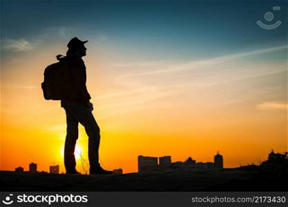 Traveler silhouette watching amazing sunset. Young casual man with backpack and cowboy hat standing alone on hill above evening cityscape. Lifestyle Travel Concept Outdoor Background. Kiev, Ukraine. Traveler silhouette watching amazing sunset