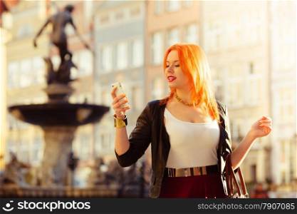 Traveler woman red hair fashion girl using cell phone smartphone outdoors in european city old town Gdansk. Neptune fountain in the background Poland