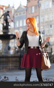 Traveler woman red haired fashion girl with smart phone outdoors in european city, old town Gdansk Neptune fountain in the background, Poland Europe