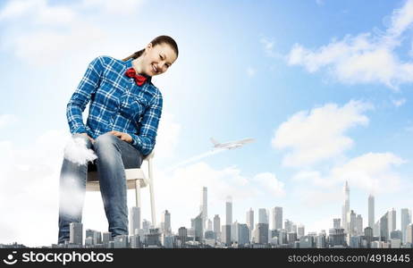 Traveling concept. Young giant woman sitting on chair above modern city