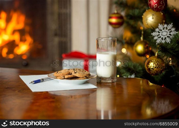 Treats and letter to Santa on wooden table next Christmas tree and burning fireplace