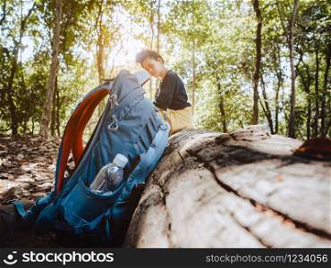 Trekking and camping adventure concept from blue backpack with tired women sitting rest on a log while walk travel nature in forest.