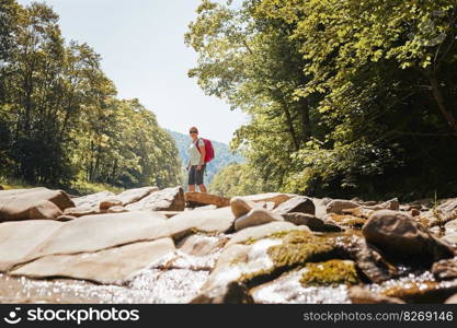 Trekking with backpack concept image. Female backpacker wearing trekking boots crossing mountain river. Woman hiking in mountains during summer trip. Vacation trip close to nature. Natural scenery