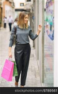 Trendy adult woman carrying paper bags and exploring display of store while walking down street in town. Content lady with shopping bags looking at showcase