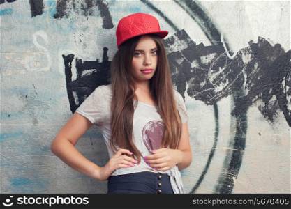 trendy beautiful long haired yuong model with big lips posing on graffiti background. Blowing bubblegum. red cap. grey t-shirt.