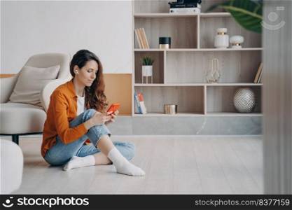 Trendy european woman is listening to music in airpods. Weekend morning relaxation. Wireless earphones. Girl sitting on floor at home downloading tracks to phone through wi-fi. Online music listening.. Trendy european woman is listening to music in airpods. Weekend morning relaxation.