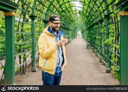 Trendy guy with beard and moustache wearing anorak, jeans and cap standing sideways having attentive look into his smartphone isolated over green arch. Human, style and modern technology concept