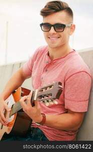 Trendy guy with guitar outdoor. Performance and show on fresh air. Young fashionable man wearing sunglasses playing classic guitar outdoor. Summer time.