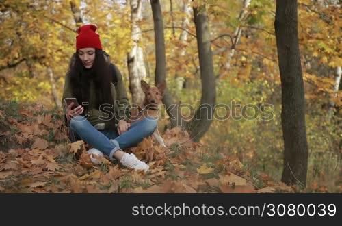 Trendy hipster girl in stylish outfit sitting on yellow fallen leaves in autumn park with her best doggy friend and surfing the net on smartphone. Woman texting on mobile phone while taking a walk with cute puppy in public park in indian summer.