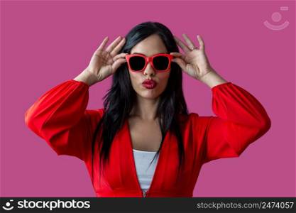 Trendy Hispanic woman in bright clothes pouting lips and adjusting sunglasses against purple background.. Stylish female putting on sunglasses