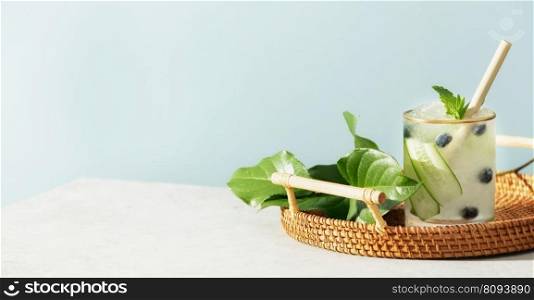 Trendy summer drink with cucumber, mint and blueberry in wicker tray on blue sky background, banner