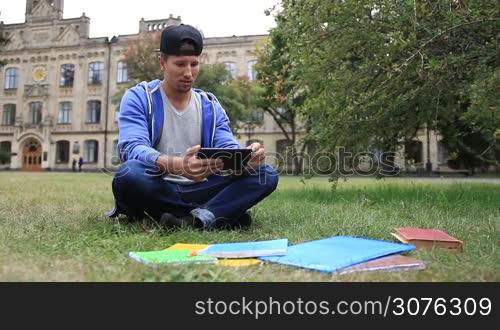 Trendy young handsome student sitting on the green grass in the park outside college and e-learning on digital tablet. Serious stylish male student studying outdoors using pc tablet.
