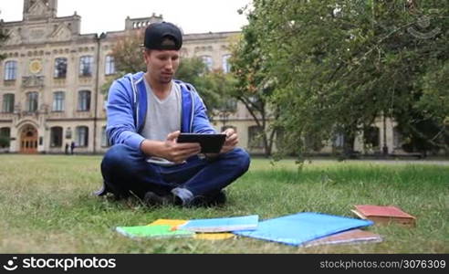 Trendy young handsome student sitting on the green grass in the park outside college and e-learning on digital tablet. Serious stylish male student studying outdoors using pc tablet.