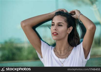 Trendy young woman in white t-shirt looking away and thinking while standing against glass wall of building and adjusting ponytail on city street. Thoughtful lady adjusting ponytail on street