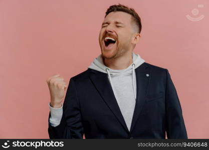 Triumphant unshaven man celebrates success, raising clenched fist, screaming in joy against pink background