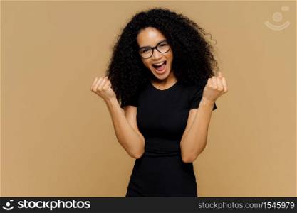 Triumphing lovely dark skinned woman raises clenched fists, yells with joy, celebrates victory, wears black clothing, stands against brown background, feels like winner. Body language, success concept