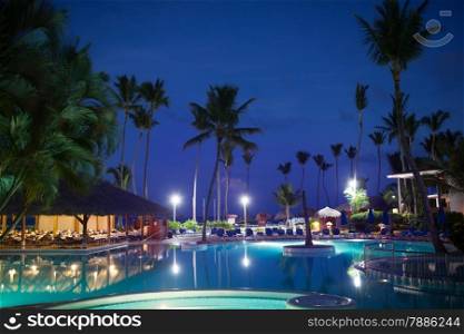 Tropical resort at night. View with empty restautant, huge palms and electric lights reflecting in the swimming pool in foreground