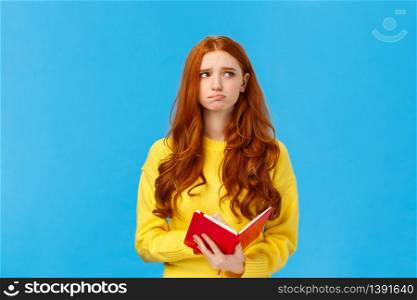 Troubled and pensive uneasy pretty redhead female, pouting and frowning looking away thoughtful, feeling sad and distressed, reading something bad in notebook, standing blue background.. Troubled and pensive uneasy pretty redhead female, pouting and frowning looking away thoughtful, feeling sad and distressed, reading something bad in notebook, standing blue background