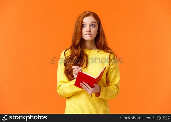 Troubled redhead caucasian girl forgot about appointment, remember task after looking notebook, biting lip and stare concerned or embarrassed, writing down important notes, orange wall.. Troubled redhead caucasian girl forgot about appointment, remember task after looking notebook, biting lip and stare concerned or embarrassed, writing down important notes, orange wall