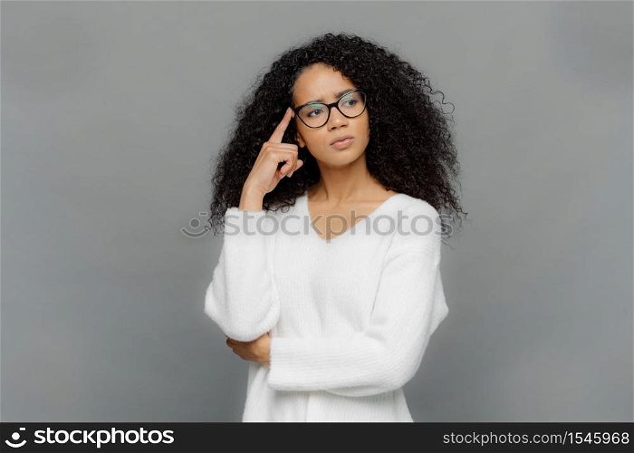 Troubled serious female keeps fore finger on temple, looks aside, has curly hair, being deep in thoughts, wears white jumper and optical glasses, isolated on grey background. People and thinking