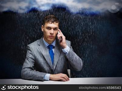 Troubles in business. Young troubled businessman sitting under rain and talking on phone