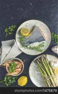 Trout fish surrounded parsley, lemon, shrimp, prawn, asparagus in ceramic plates. Black concrete table surface. Healthy seafood background.