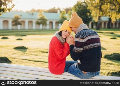 True feelings and romantisism concept. Adorable young woman in knitted yellow hat and red warm sweater warms her hands in boyfriend s hands, sit together on bench, closes eyes with great enjoyment