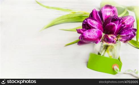 Tulips bunch in glass jar with water and empty tag on light background, banner. Spring flowers concept.