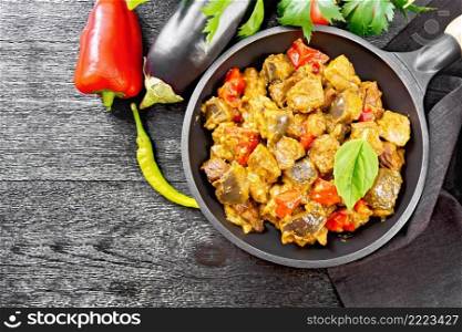 Turkey or pork meat stew with eggplant, onion, tomato and pepper in a frying pan, towel, basil and parsley on wooden board background from above