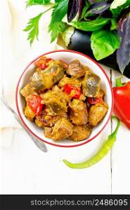 Turkey or pork meat stew with eggplant, onion, tomato and pepper in a bowl, napkin, basil and parsley on light wooden board background from above