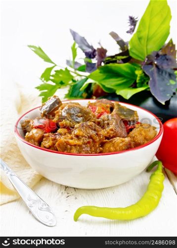 Turkey or pork meat stew with eggplant, onion, tomato and pepper in a bowl, towel, basil and parsley on light wooden board background