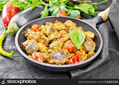 Turkey or pork meat stew with eggplant, onion, tomato and pepper in a frying pan, towel, basil and parsley on wooden board background