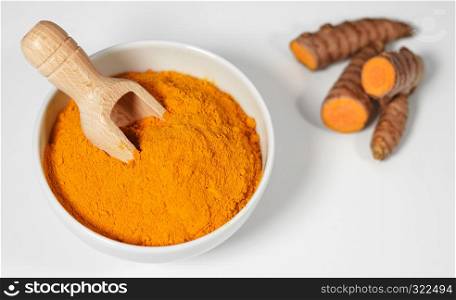 Turmeric powder and roots healthy spice Asian food with sliced turmeric roots and powder in a bowl with a wooden bailer on white background.