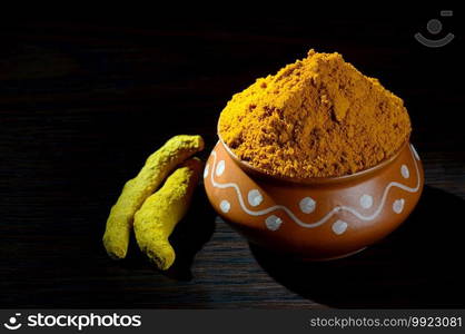 Turmeric powder in clay pot with roots or barks on black background