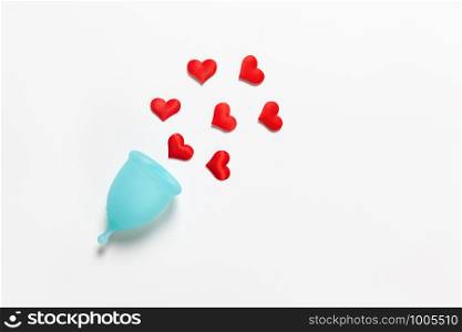 Turquoise menstrual cup on white background with red hearts. Concept zero waste, savings, minimalism, these days. Feminine hygiene product, flat lay, copy space. Horizontal.