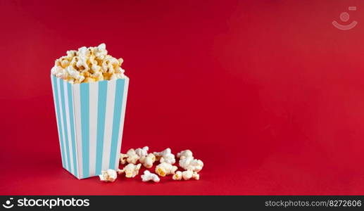 Turquoise white striped carton bucket with tasty cheese popcorn, isolated on red background. Box with scattering of popcorn grains. Fast food, movies, cinema and entertainment concept.