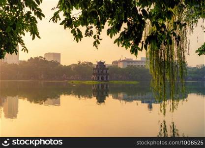 Turtle tower with modern skyscraper buildings and reflection in the middle of Hoan Kiem Lake in public park garden with trees in Downtown Hanoi. Urban city at sunset, Vietnam. Cityscape background.