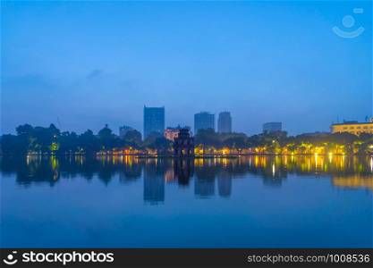 Turtle tower with modern skyscraper buildings and reflection in the middle of Hoan Kiem Lake in public park garden with trees in Downtown Hanoi. Urban city at sunset, Vietnam. Cityscape background.