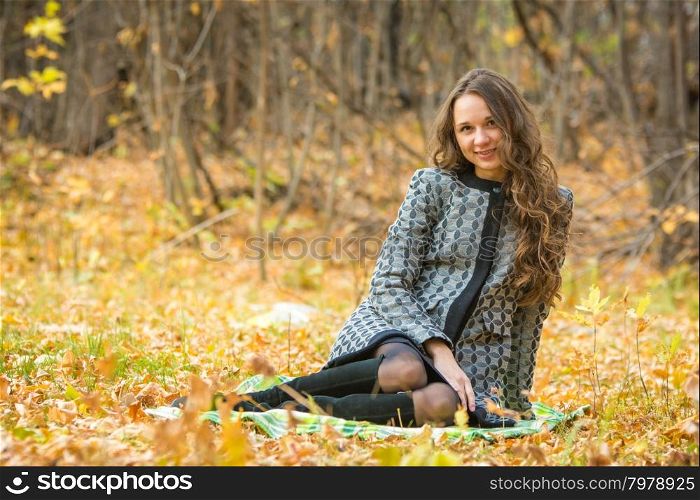 Twenty-five young beautiful girl walks by Europeans autumn forest. Young beautiful girl in a yellow coat sitting on fallen leaves in the forest