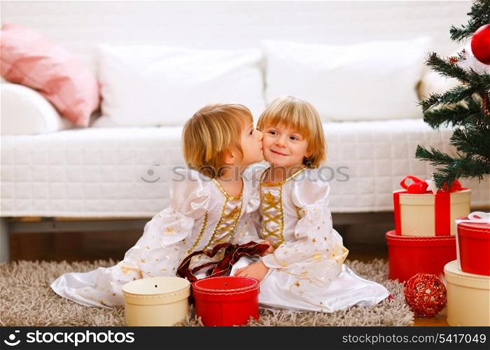 Twin girl kissing her sister near Christmas tree with gifts&#xA;