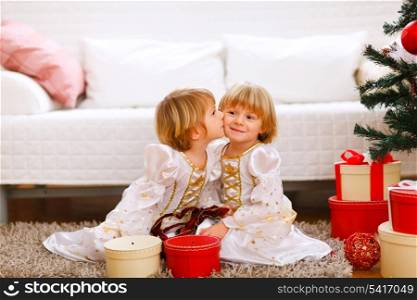 Twin girl kissing her sister near Christmas tree with gifts&#xA;