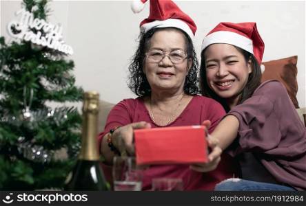 Two 60s old mother and daughter wearing santa hat, hug and holding gift at home with blur foreground of tree and bottle. Christmas or New Year Celebration Concept.